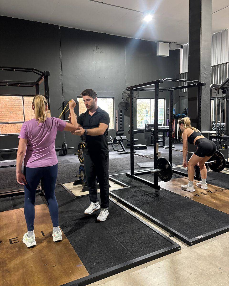 Max giving hands-on feedback during a banded shoulder exercise while another client deadlifts behind