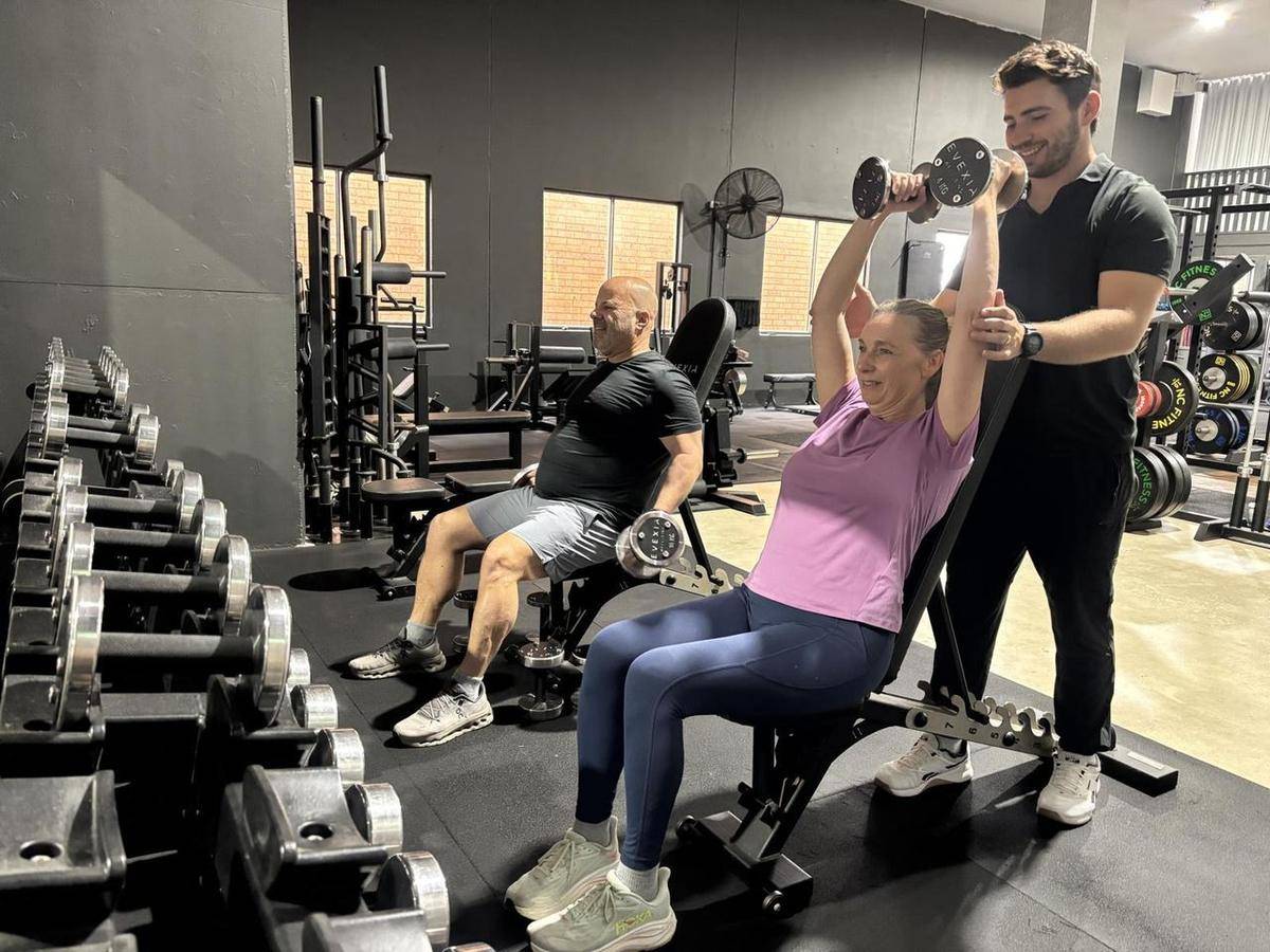 A client in her 40s pressing dumbbells overhead with Max spotting
